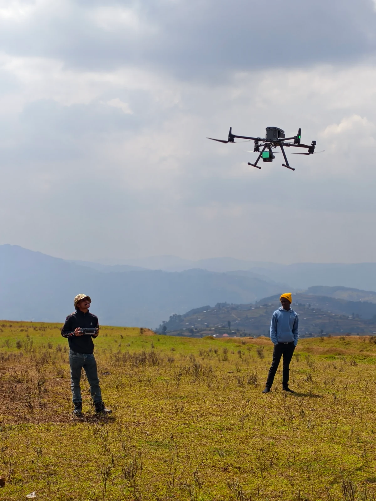 Aircraft in flight above a grassy Uganda field, with two people operating from the ground
